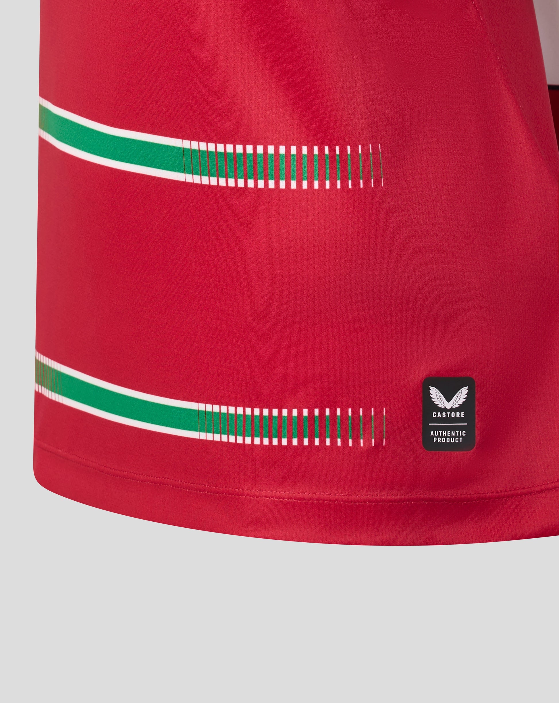 Close-up of a red athletic jersey or shorts fabric with two horizontal green and white stripe accents, a dashed green stripe detail, and a small black Wings brand patch near the lower right.