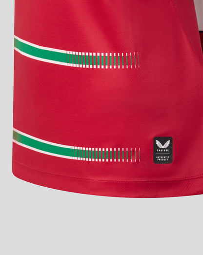 Close-up of a red athletic jersey or shorts fabric with two horizontal green and white stripe accents, a dashed green stripe detail, and a small black Wings brand patch near the lower right.