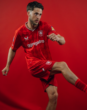 Male model wearing FC Twente 25/26 home shirt in an action pose against a red background
