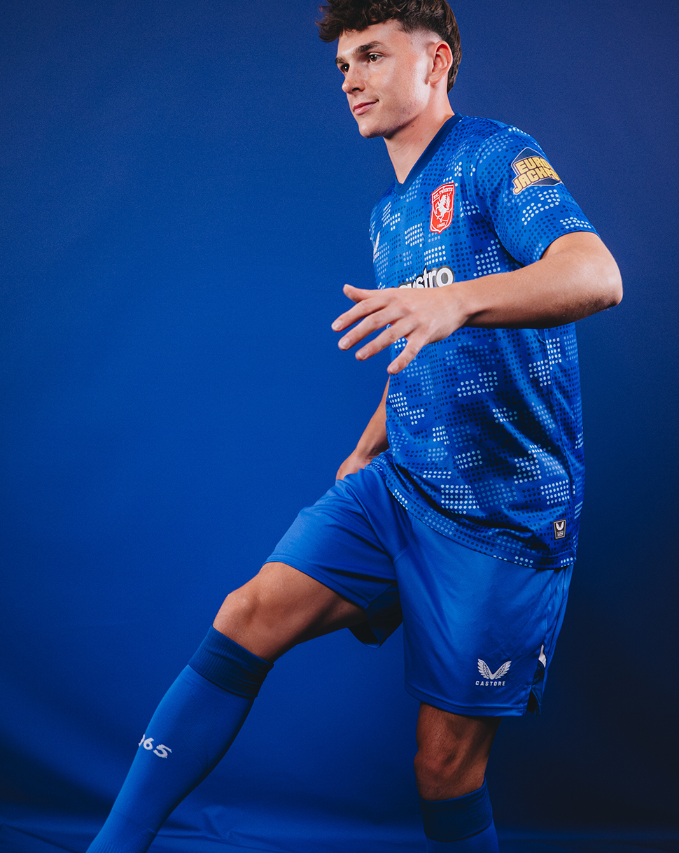 A male soccer player in a blue patterned jersey, matching shorts, and blue socks, posed dynamically against a solid blue studio background.