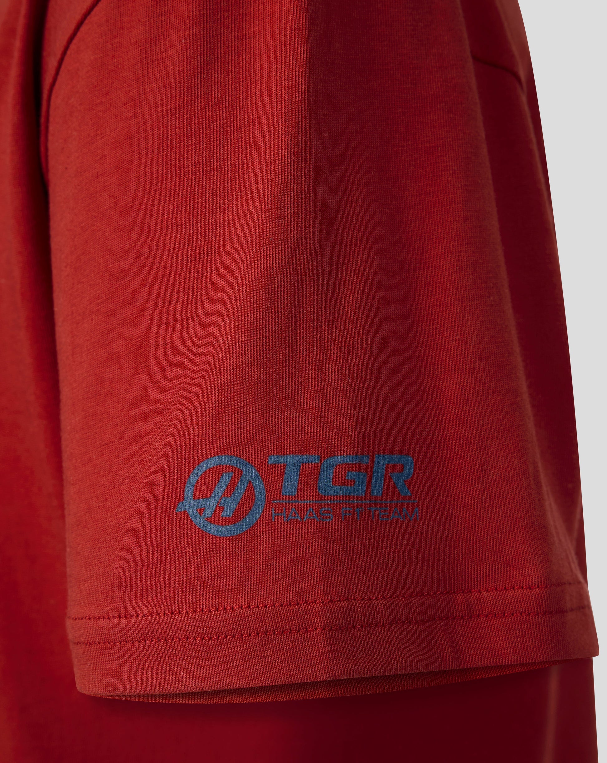 Close-up of a red short-sleeve t-shirt showing the left sleeve with a blue TGR Apparel logo printed on the fabric.