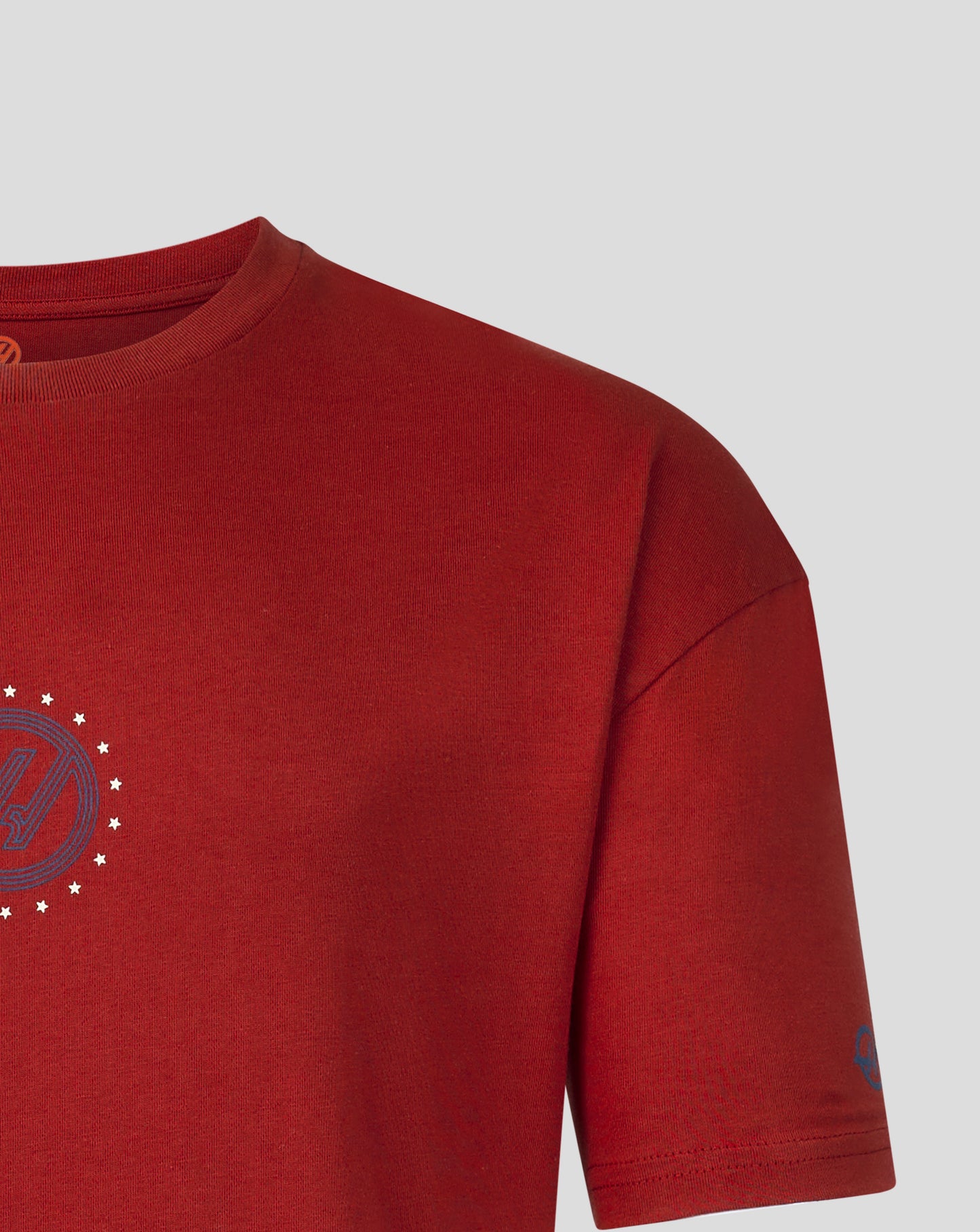 A close-up of a red short-sleeve t-shirt showing the chest area with a circular graphic on the left side and a small blue graphic on the sleeve, set against a light gray background.