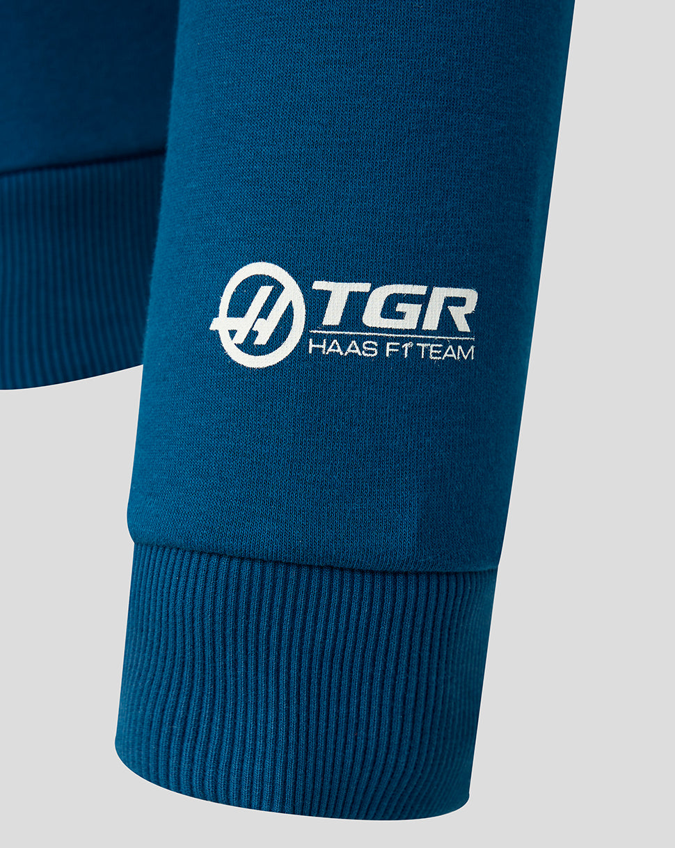 Close-up of a blue ribbed sweatshirt cuff featuring a white printed logo that reads 'TGR HAAS FT TEAM' with a circular interlocking H emblem.
