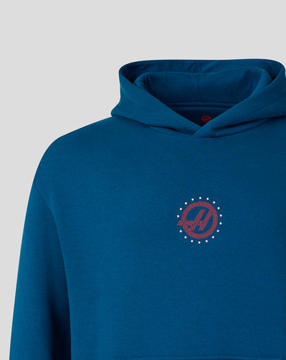 A close-up of a blue hoodie featuring a red circular chest graphic with a stylized 'H' inside a diagonal slash, surrounded by a ring of small white dots, on a light gray background.