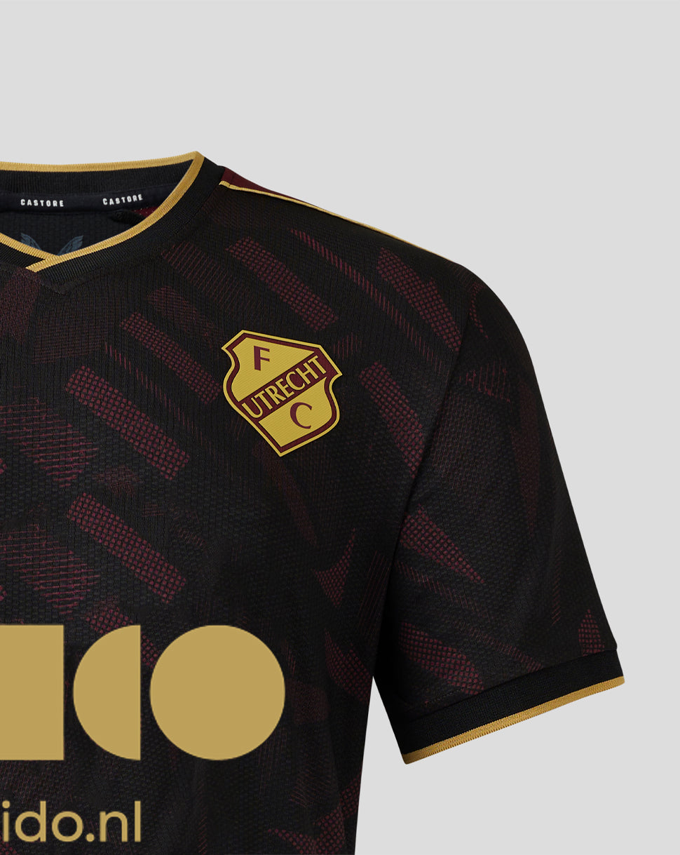 Shoulder and chest close up of FC Utrecht junior away shirt with crest on the left and gold trim on the sleeve against the textured pattern