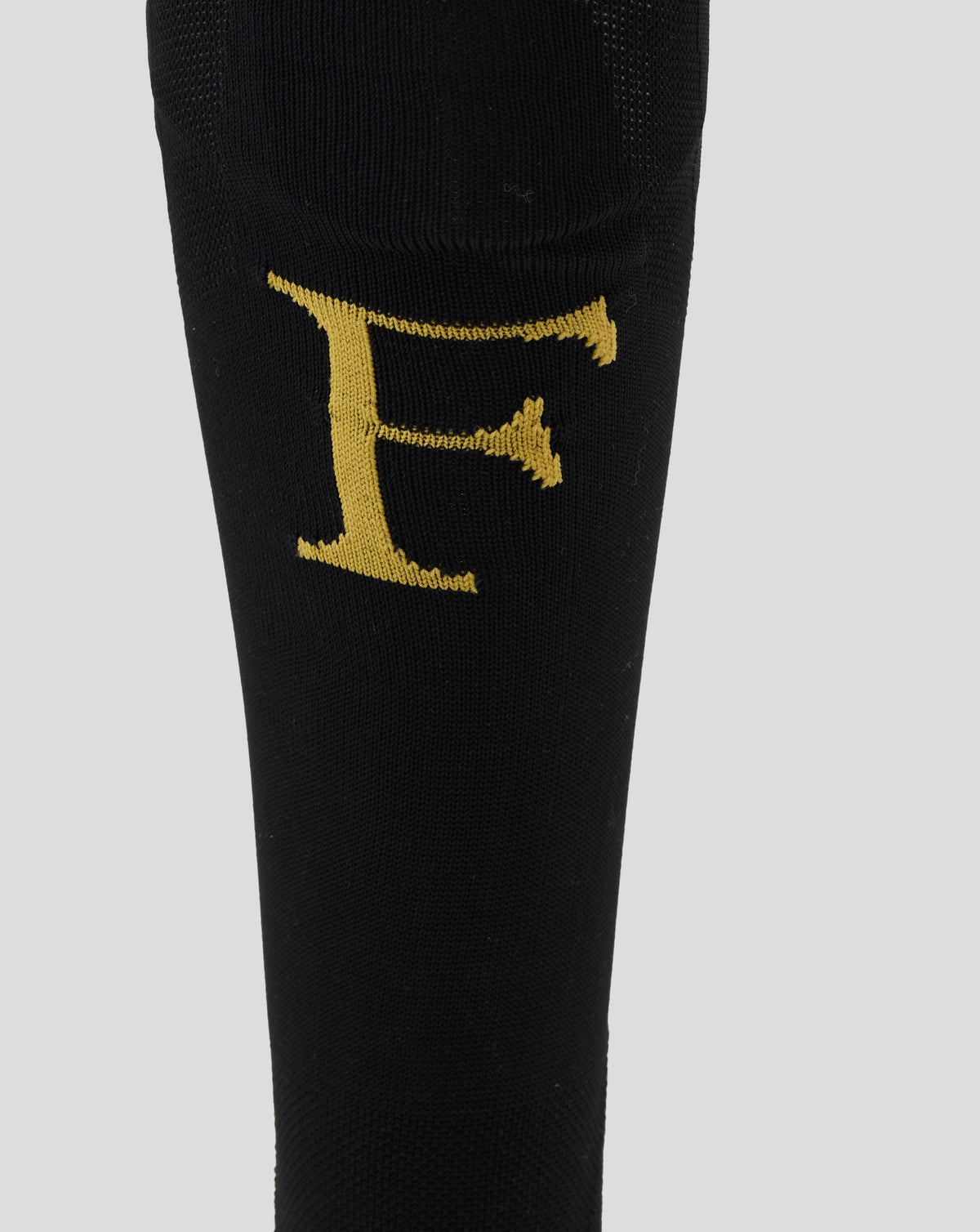 A close-up of a black knit sock featuring a large, textured gold letter 'F' on the side, set against a solid light gray background.
