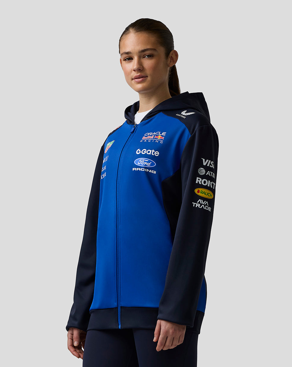 Person wearing a blue and black jacket with various logos on a gray background