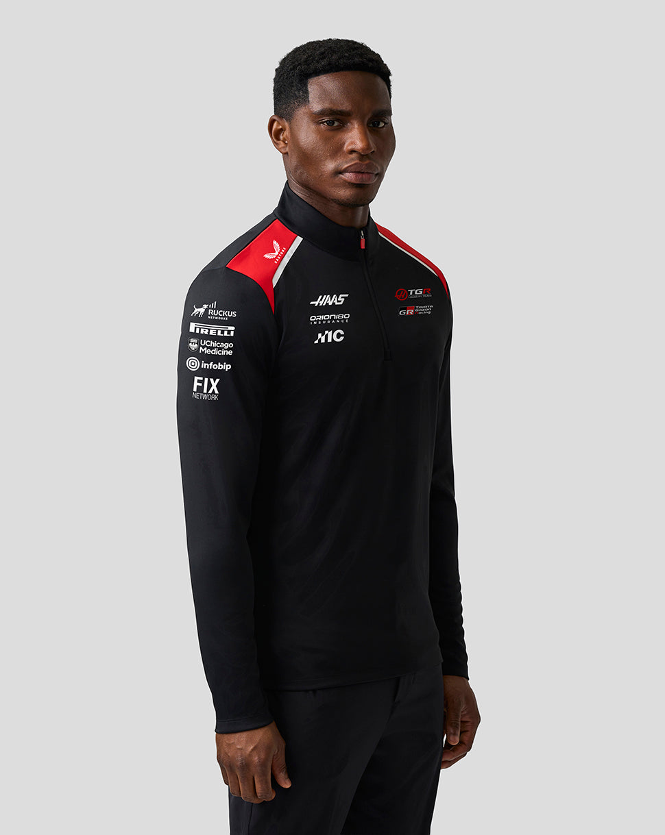 Person wearing a black tracksuit with various logos on a gray background