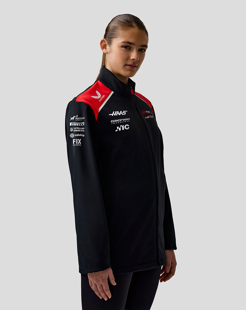 Person wearing a black jacket with sponsor logos on a gray background