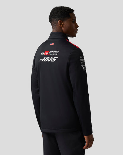 Person wearing a black jacket with sponsor logos on a gray background