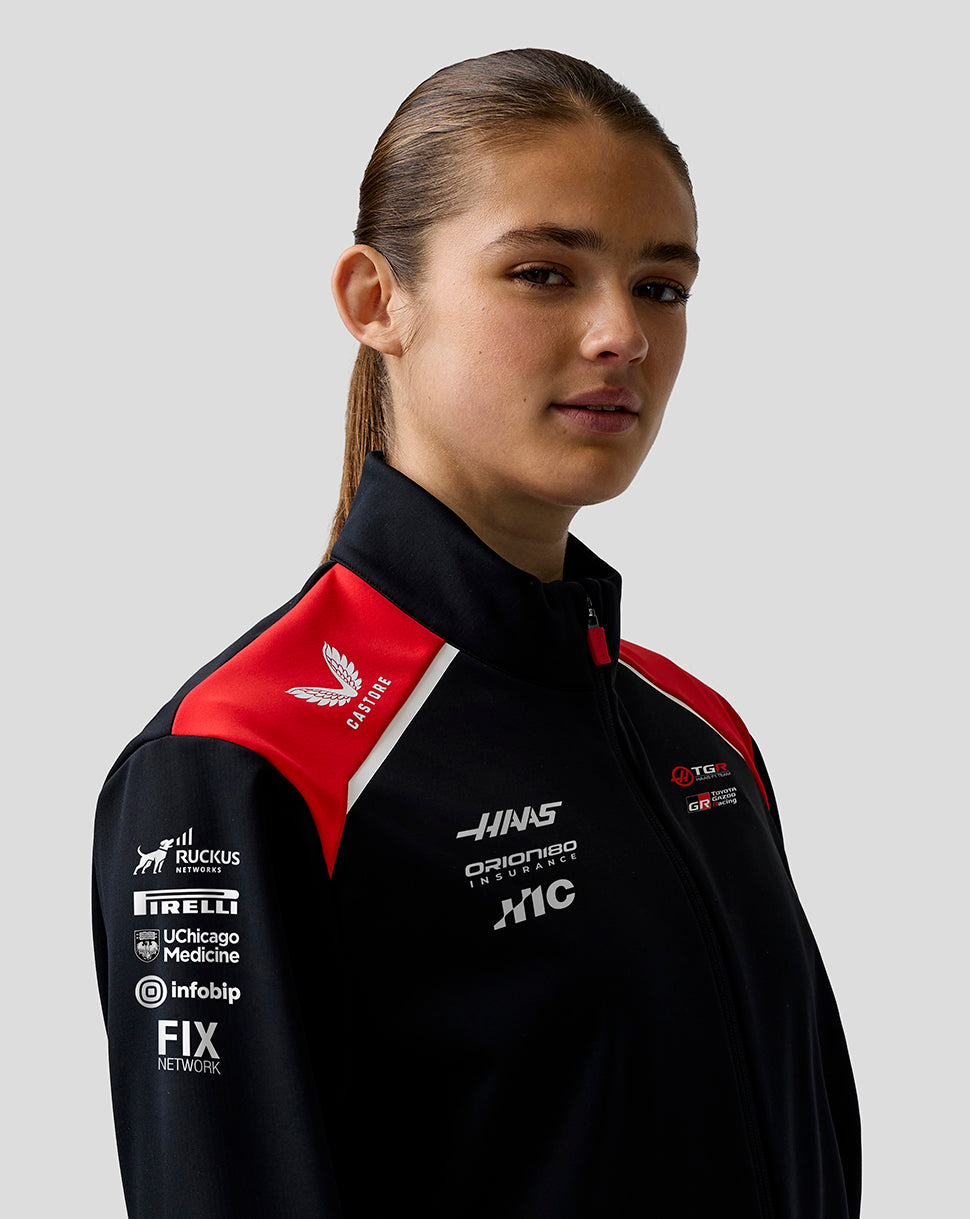 Person wearing a black jacket with red and white accents featuring various logos on a gray background