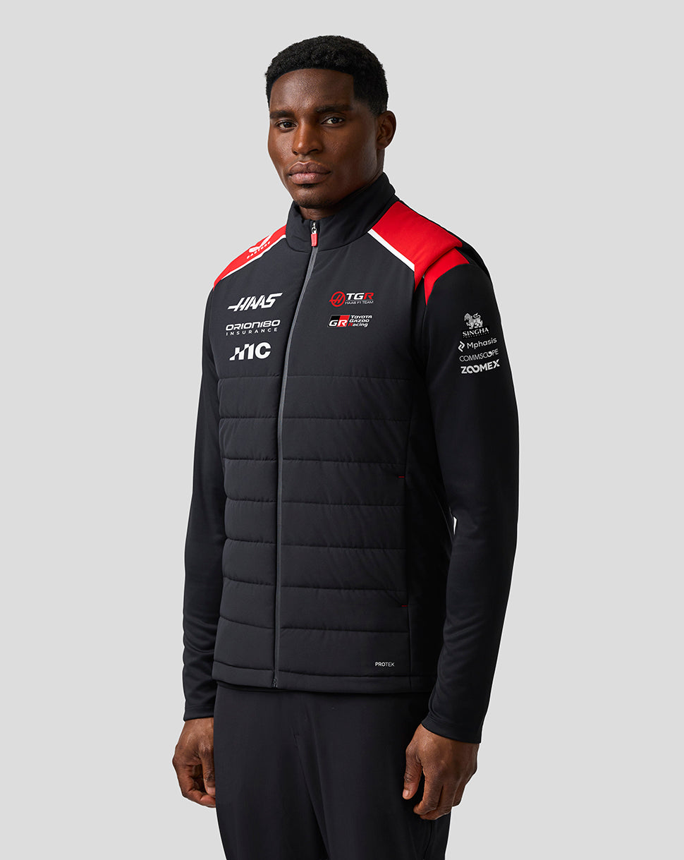 Man wearing a black jacket with various logos on a gray background