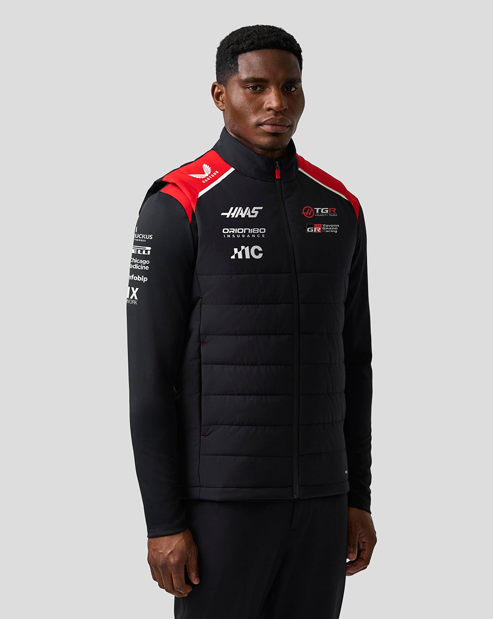 Person wearing a black puffer jacket with sponsor logos on a gray background