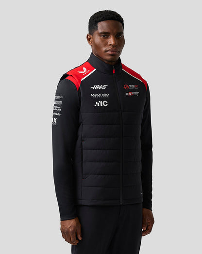 Person wearing a black puffer jacket with sponsor logos on a gray background