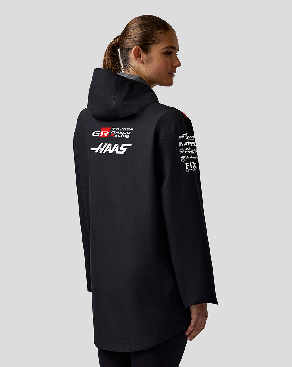 Person wearing a black jacket with various logos on the back against a light gray background