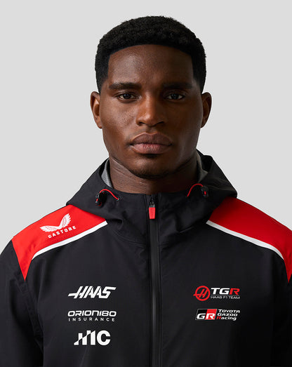 Person wearing a black jacket with multiple logos on a gray background