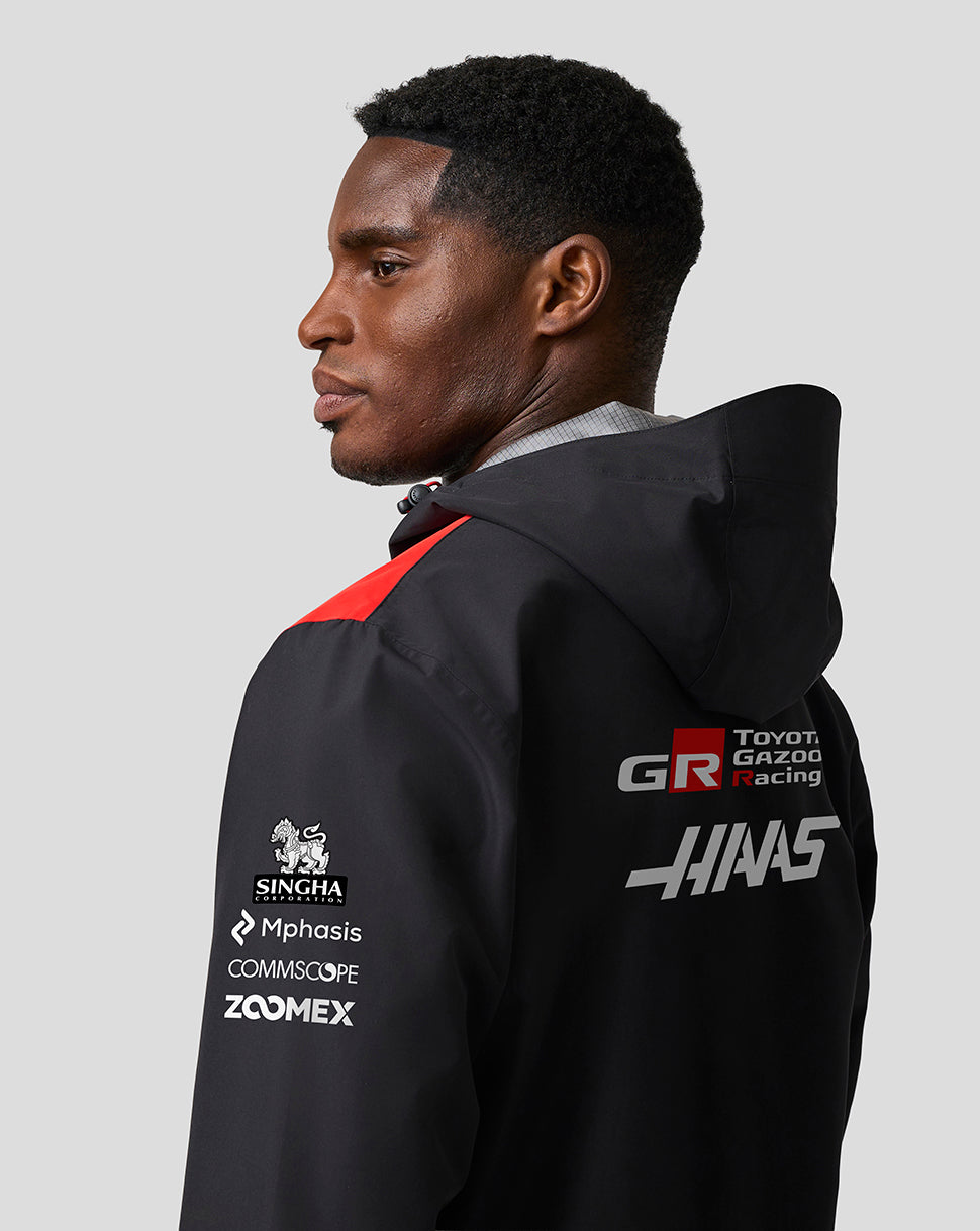 Person wearing a black jacket with various logos on the back against a gray background