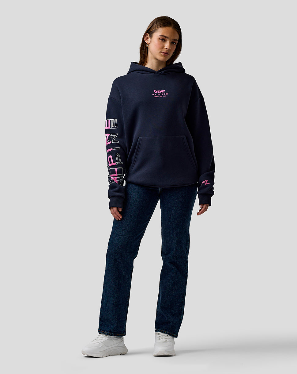 Female model in navy BWT Alpine graphic hoodie with pink chest logo and bold sleeve artwork, standing against a gray backdrop.