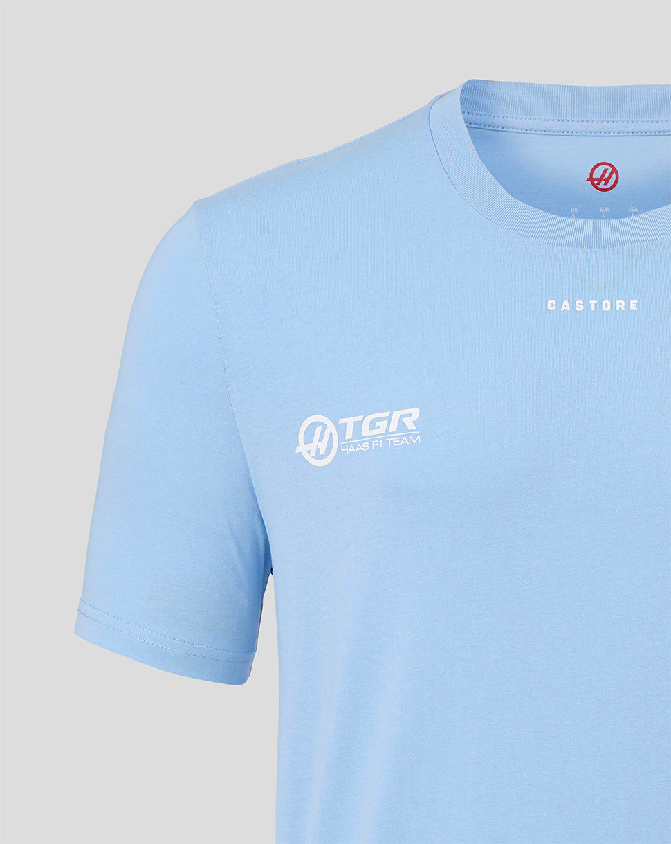 A close-up of a light blue athletic t-shirt featuring white printed logos on the chest, including 'TGR' and 'CASTORE', on a light gray background.